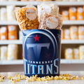 A tin with the Tennessee Titans logo, with bags of Marigold popcorn inside and sitting on the side. Loose popcorn strewn about the base.