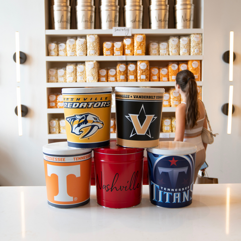 Four sports tins, stacked, in the foreground, representing the Nashville Predators, Vanderbilt University, the Tennessee Vols, the Tennessee Titans, and the City of Nashville. A person with a pony tail shops popcorn in the background.