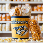 A tin with the Nashville Predators logo, with bags of Marigold popcorn inside and sitting on the side. Loose popcorn strewn about the base. 