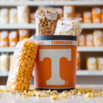 A tin with the Tennessee Vols logo, with bags of Marigold popcorn inside and sitting on the side. Loose popcorn strewn about the base. 