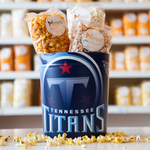 A tin with the Tennessee Titans logo, with bags of Marigold popcorn inside and sitting on the side. Loose popcorn strewn about the base. 