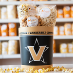 A tin with the Vanderbilt University logo, with bags of Marigold popcorn inside and sitting on the side. Loose popcorn strewn about the base. 