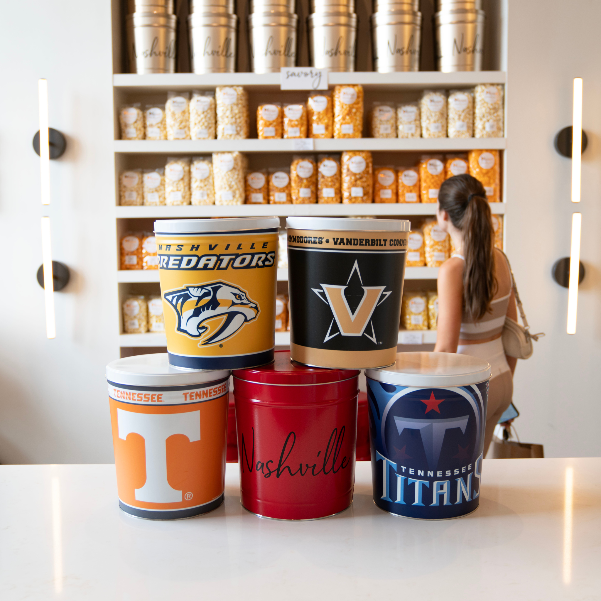 Four sports tins, stacked, in the foreground, representing the Nashville Predators, Vanderbilt University, the Tennessee Vols, the Tennessee Titans, and the City of Nashville. A person with a pony tail shops popcorn in the background. 